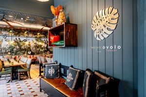 Cozy restaurant corner with a leather bench, patterned cushions, a teal wall with a monstera leaf sculpture and tropical mural.