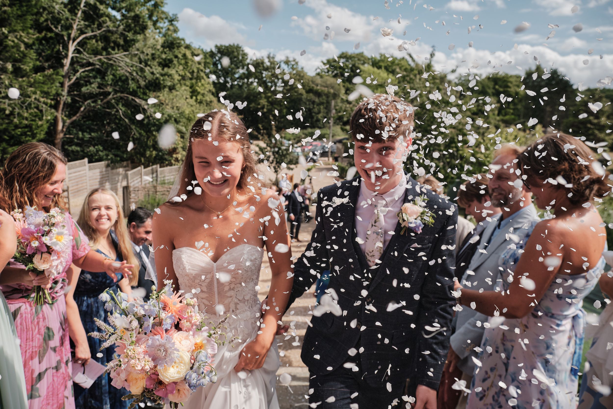 Bride and groom walk through confetti shower surrounded by cheering guests at outdoor wedding ceremony.