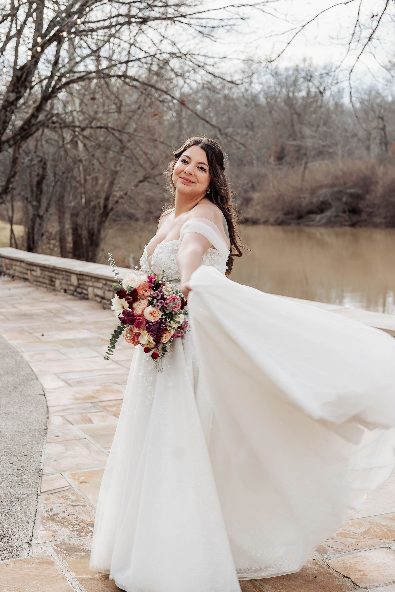 Smiling bride in flowing white off-shoulder gown holding colorful bouquet, posing by stone bridge near lake.