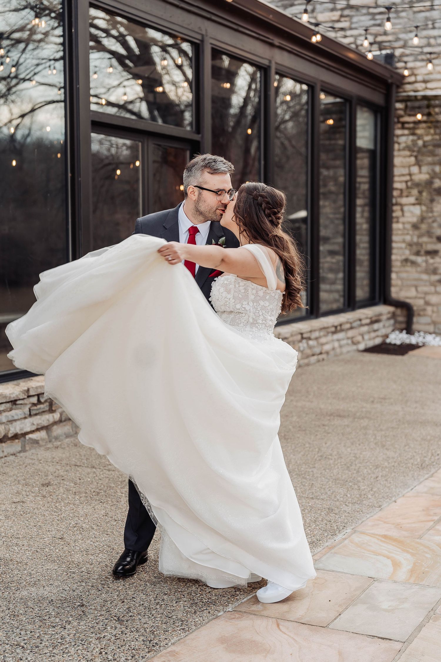 Groom lifts bride in flowing white wedding gown outdoors, sharing a romantic kiss near a stone building with string lights.