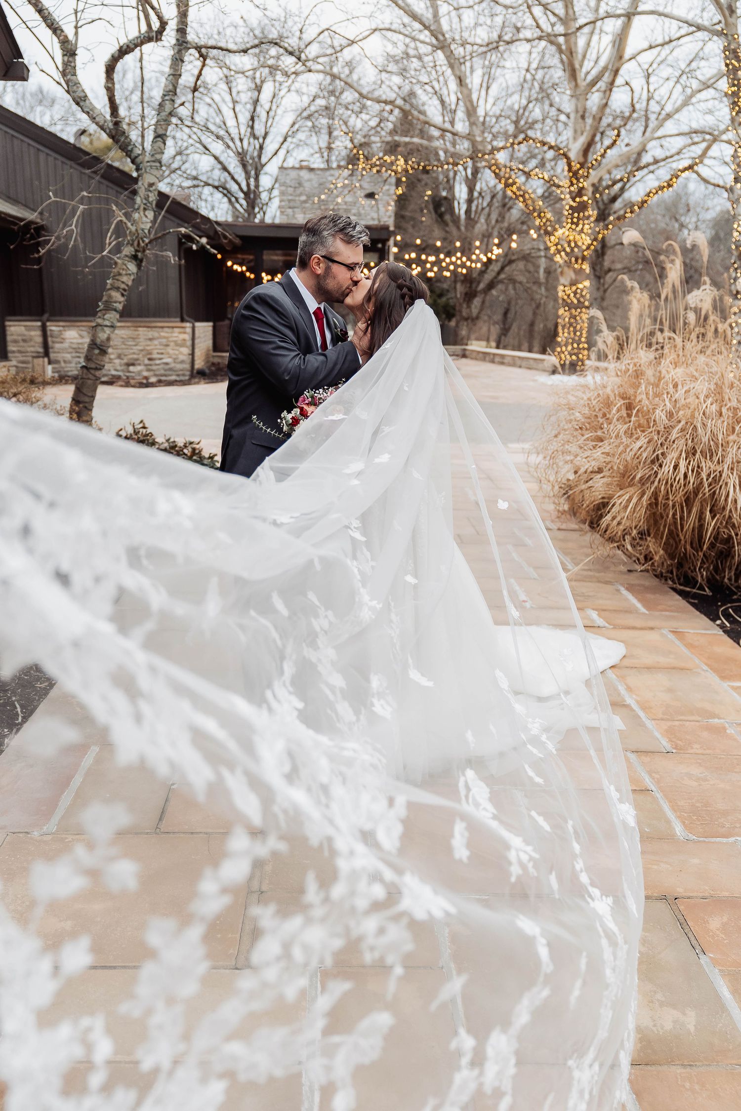 Bride and groom kissing outdoors with flowing lace veil, winter wedding venue with string lights.