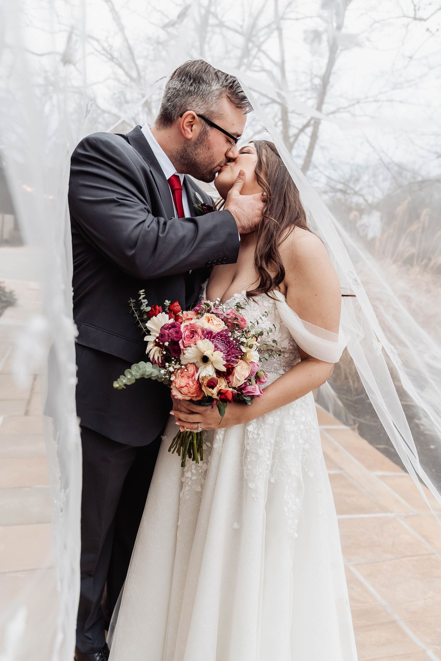 Bride and groom share a romantic kiss under a flowing veil, bride holding a pink and red floral bouquet.