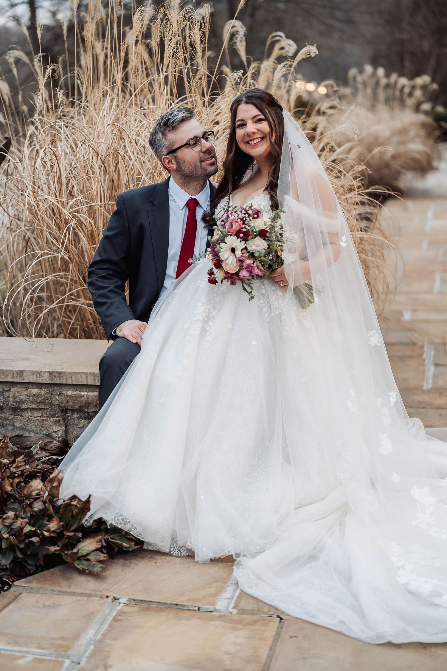 A bride in a white ball gown and groom in a dark suit pose together outdoors amid tall golden grasses.