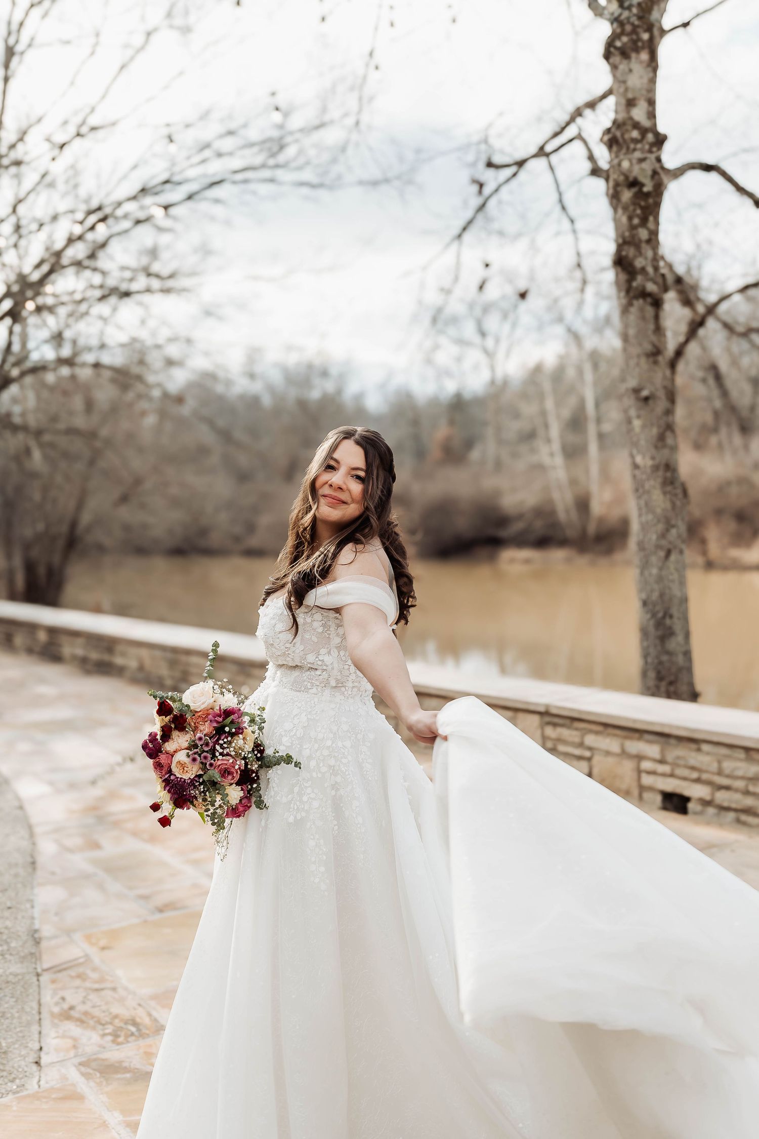 Bride in off-shoulder white gown twirling with bouquet on stone terrace near river.