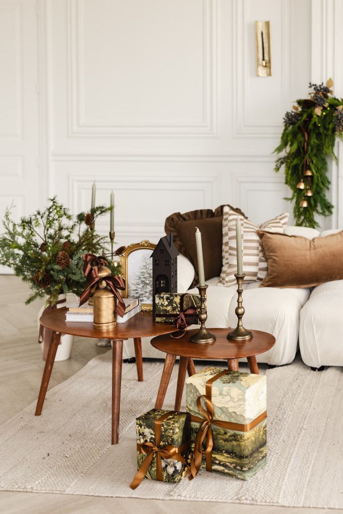 Festive living room with wooden nesting tables, brass candlesticks, wrapped gifts, evergreen garland and a cream sofa with cozy pillows.