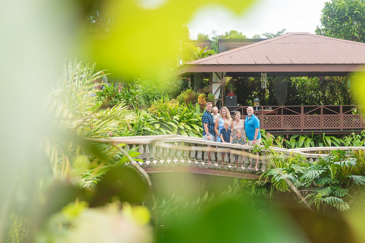 Group of people standing on a white railed bridge surrounded by lush tropical plants near a covered wooden pavilion.