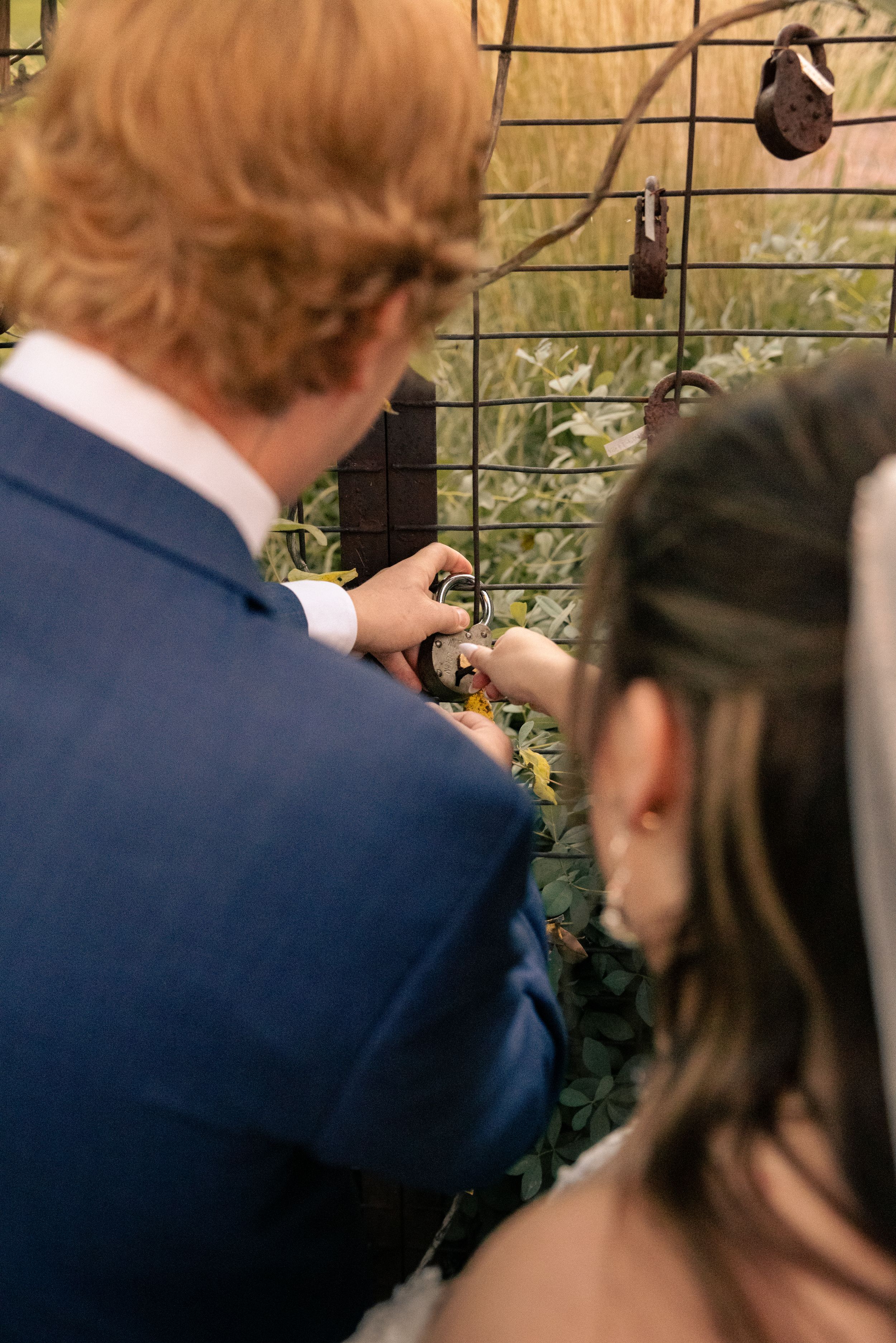 A couple attaches a love lock to a decorative wire fence at their wedding ceremony outdoors.