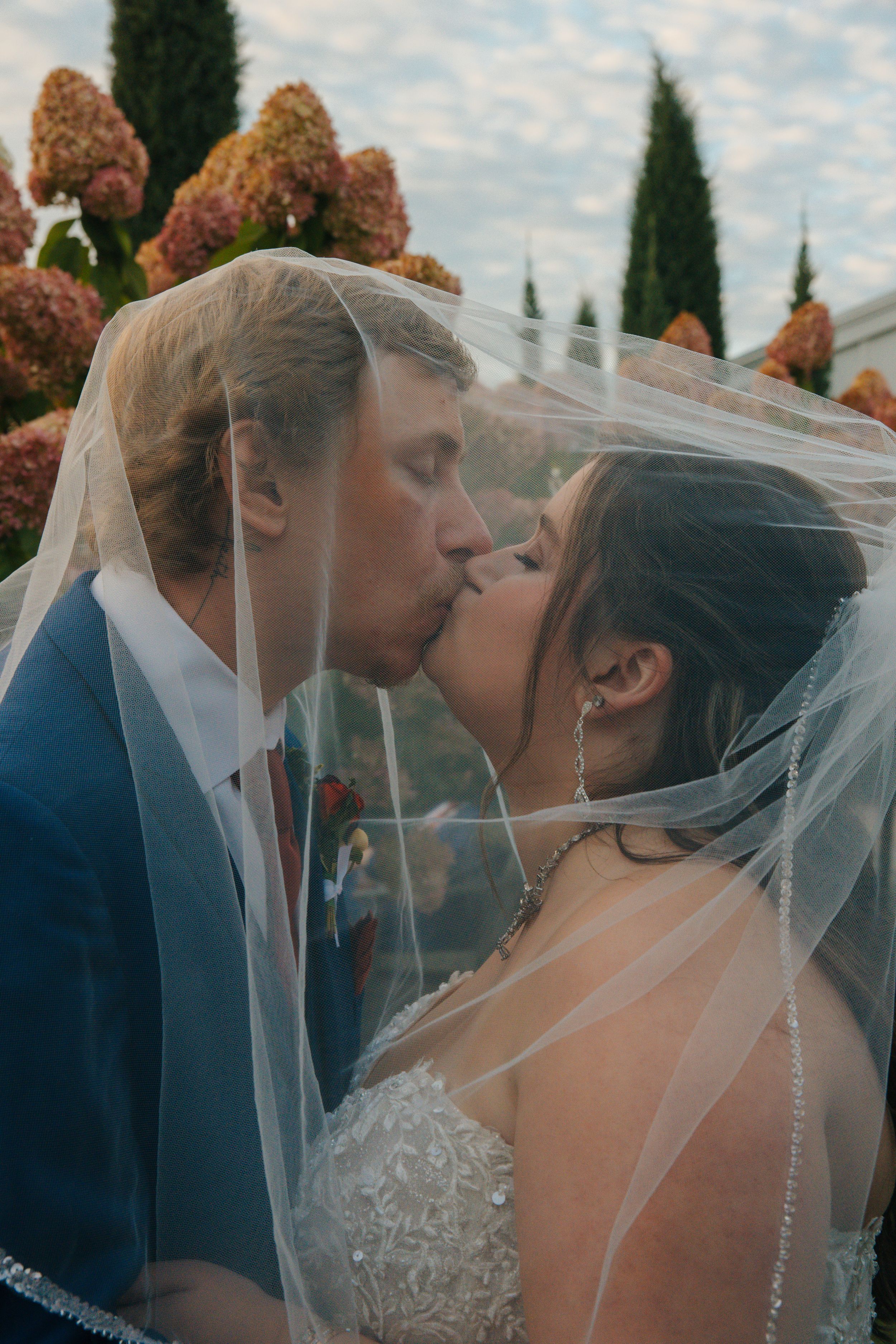 Bride and groom sharing a romantic kiss under a flowing veil, surrounded by hydrangeas and cypress trees.
