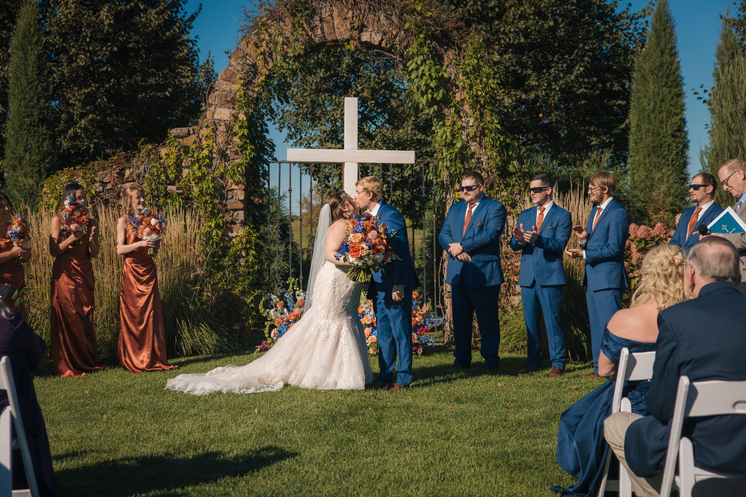 Bride and groom share first kiss at outdoor wedding ceremony with white cross arch and wedding party in blue and burgundy.