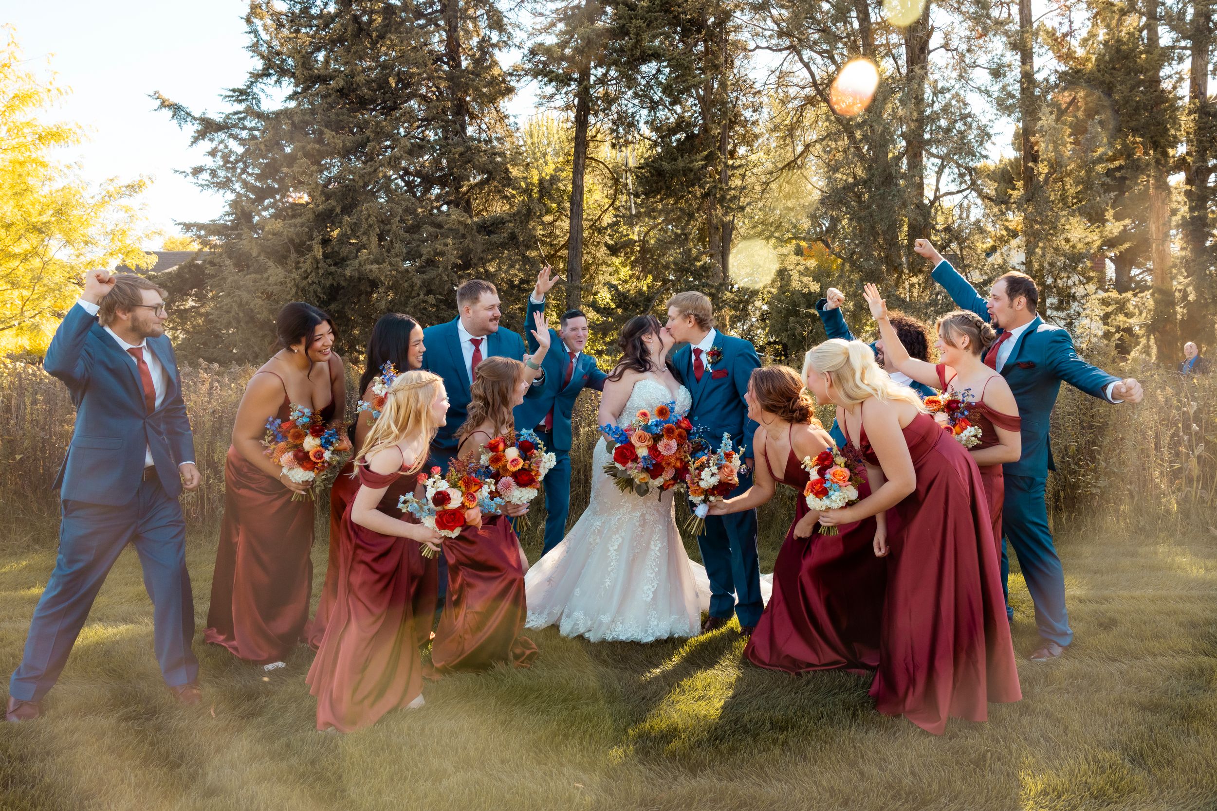 Wedding party celebrates outdoors in autumn, bridesmaids in burgundy gowns, groomsmen in blue suits, surrounded by golden trees.