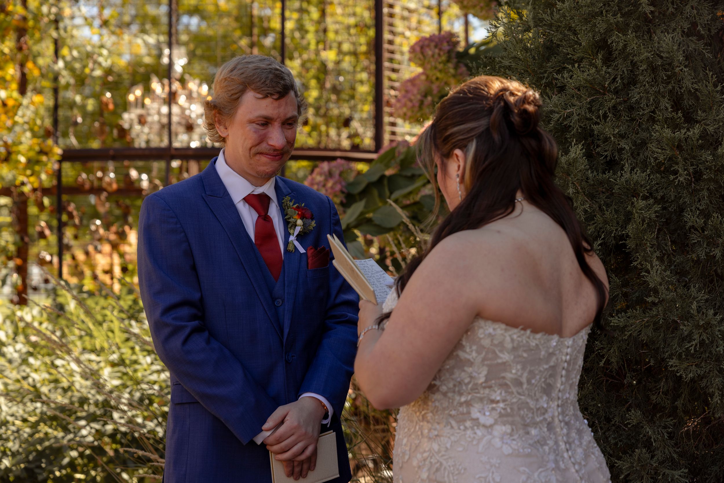 Groom in blue suit with red tie exchanges vows with bride in white lace gown during intimate outdoor wedding ceremony.