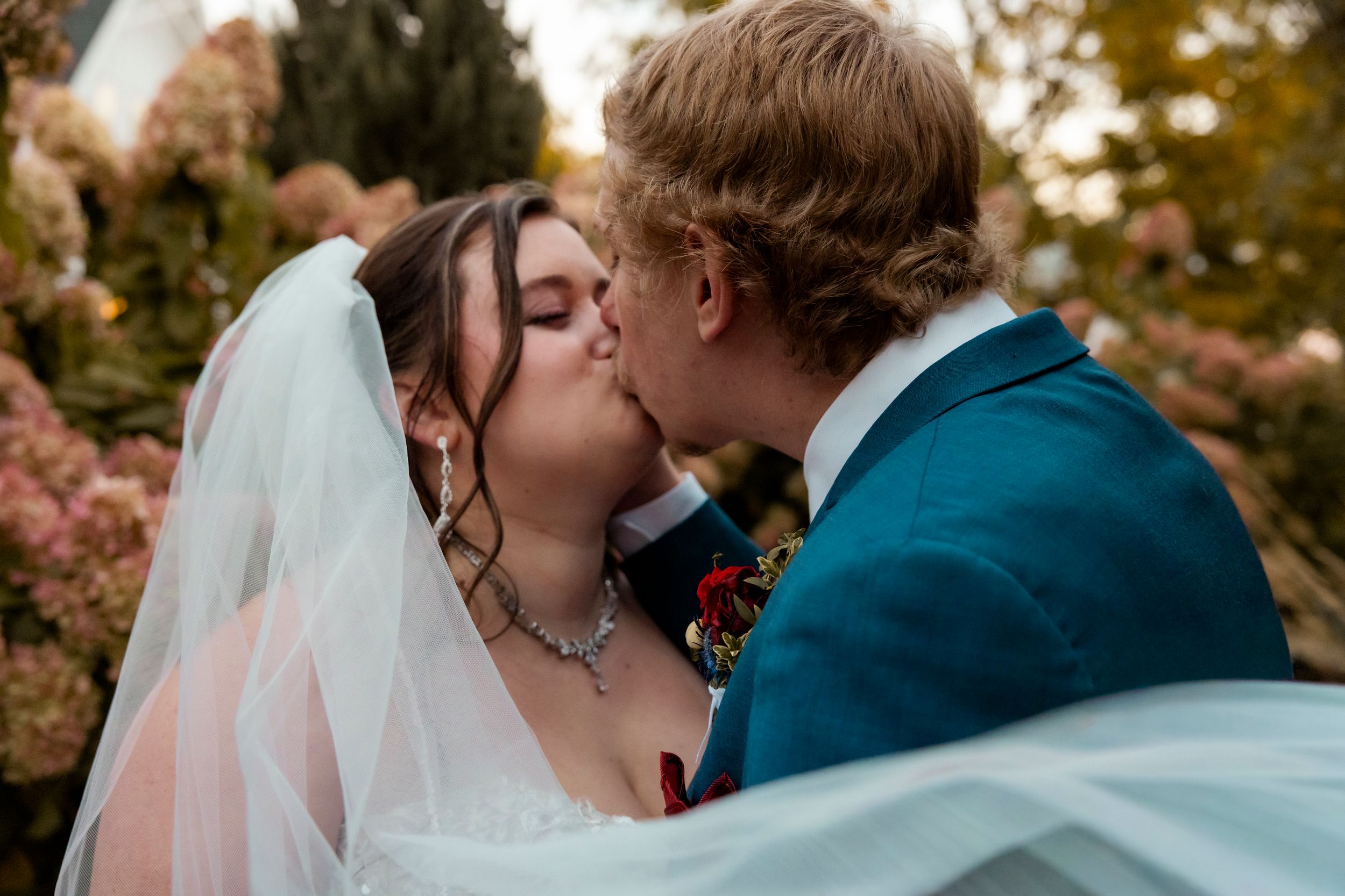 Bride and groom share a romantic first kiss, bride wearing veil and necklace, groom in teal suit, surrounded by blooming flowers.