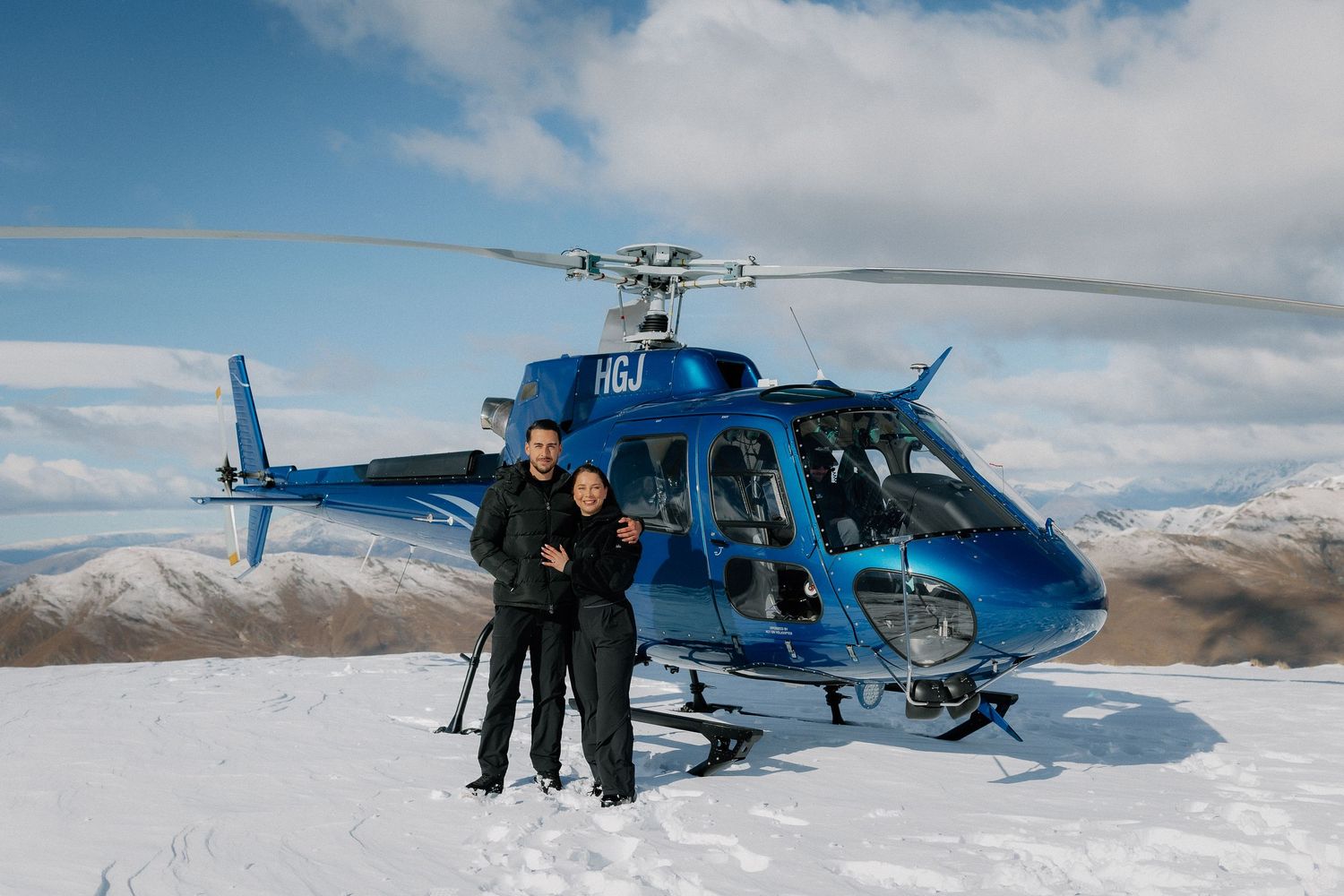 Couple posing in front of a blue helicopter on a snowy mountain summit under a partly cloudy sky.