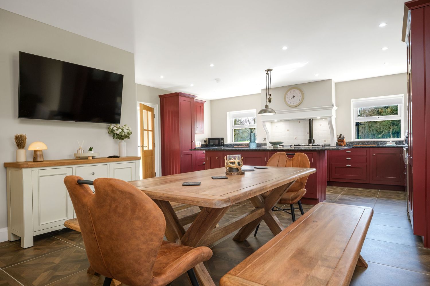 Rustic kitchen-diner with farmhouse table, leather chairs, red cabinetry, and wall-mounted television.