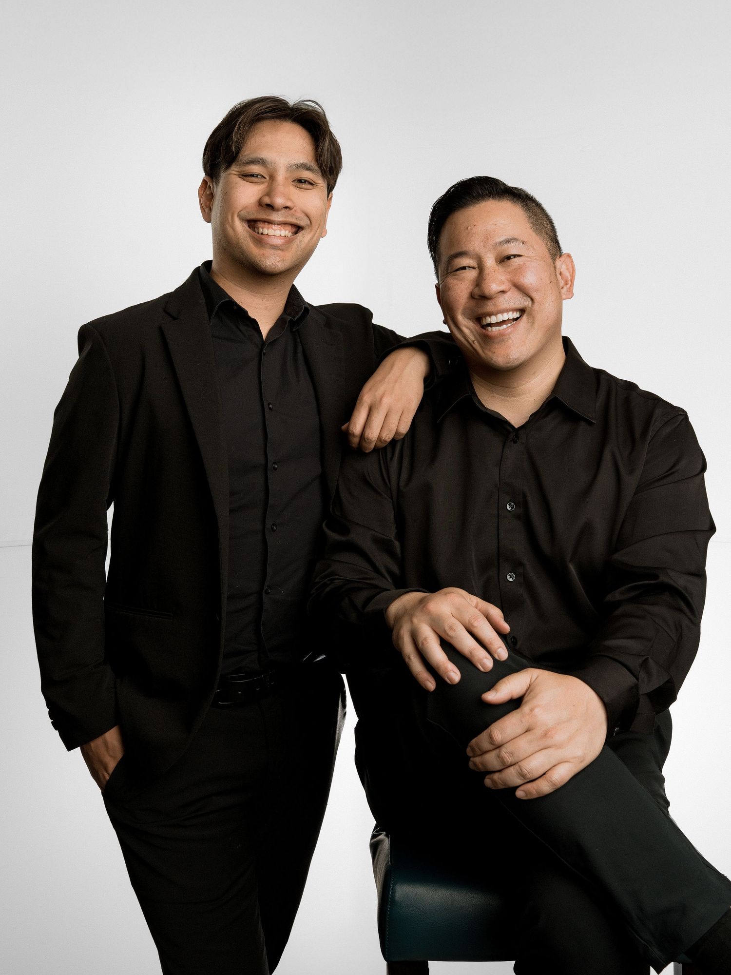 Two smiling men in black outfits pose together in a professional studio portrait against a gray background.