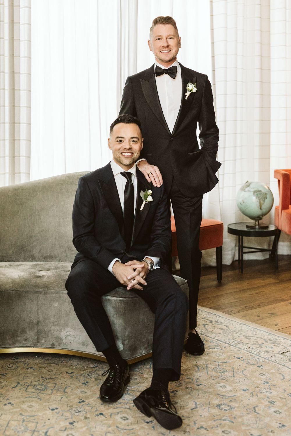Two grooms in black tuxedos pose together, one seated and one standing, wearing boutonnieres in an elegant room.