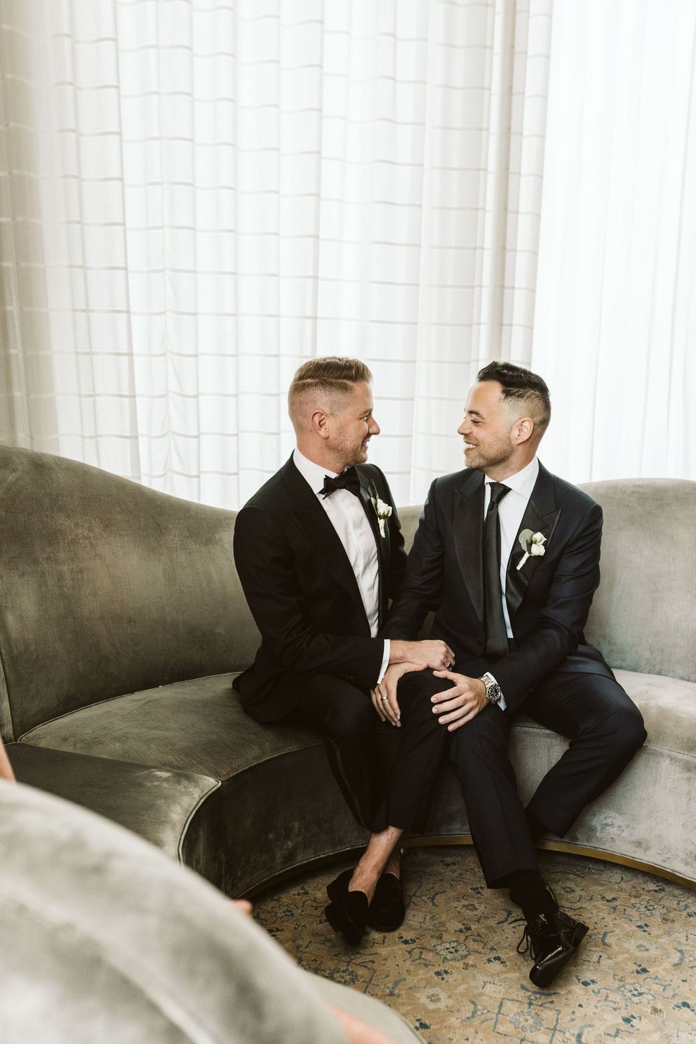 Two grooms in suits sit together on a velvet sofa, smiling at each other on their wedding day.