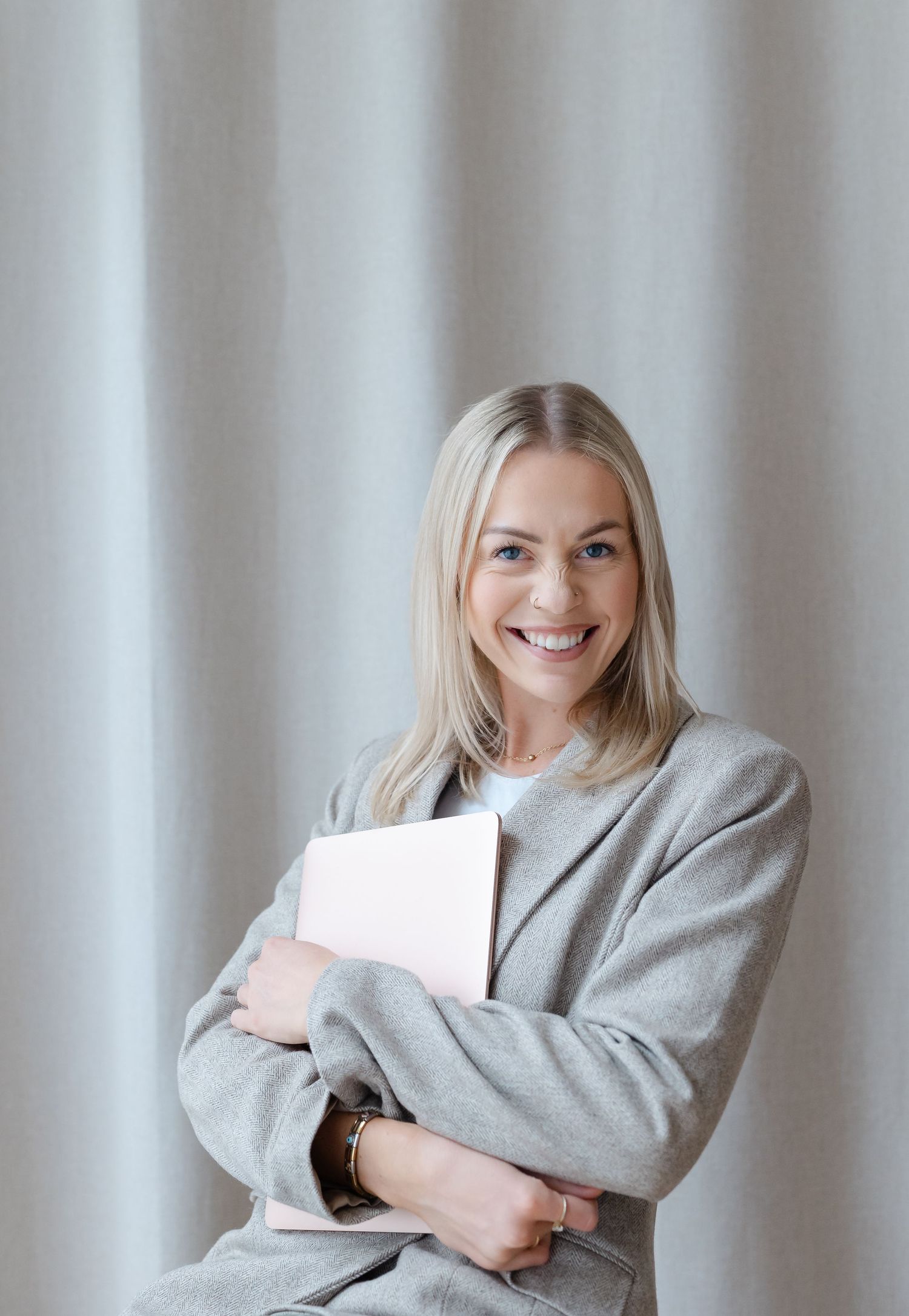 Smiling blonde woman in gray blazer holding a laptop, standing against a light curtain background.