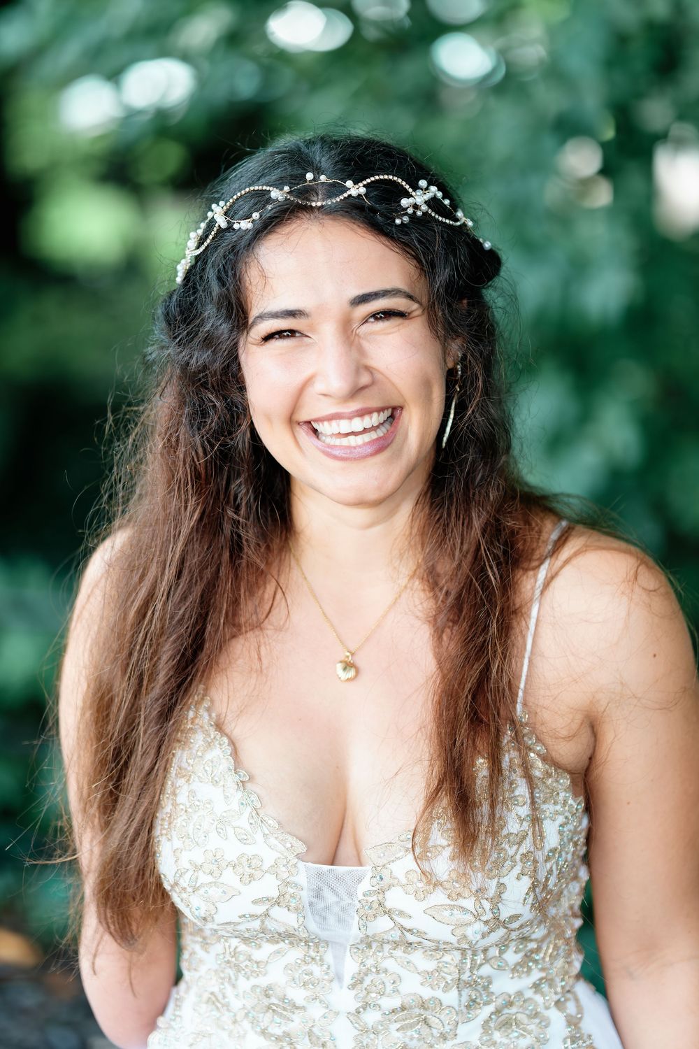 Smiling bride wearing floral crown and white lace dress laughing outdoors with green foliage background.