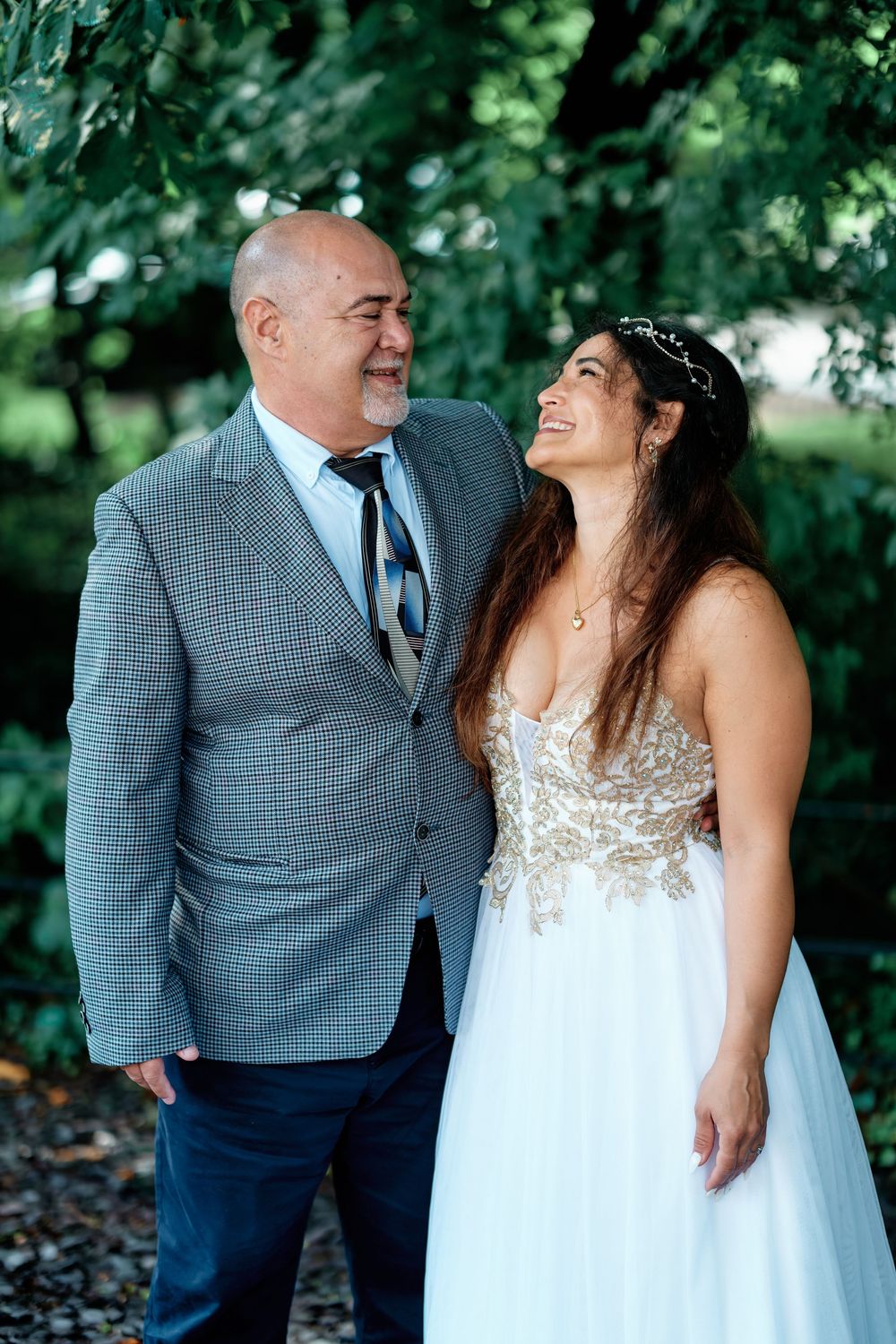 A bride in a white lace gown and groom in a gray suit share a joyful moment outdoors surrounded by lush greenery.