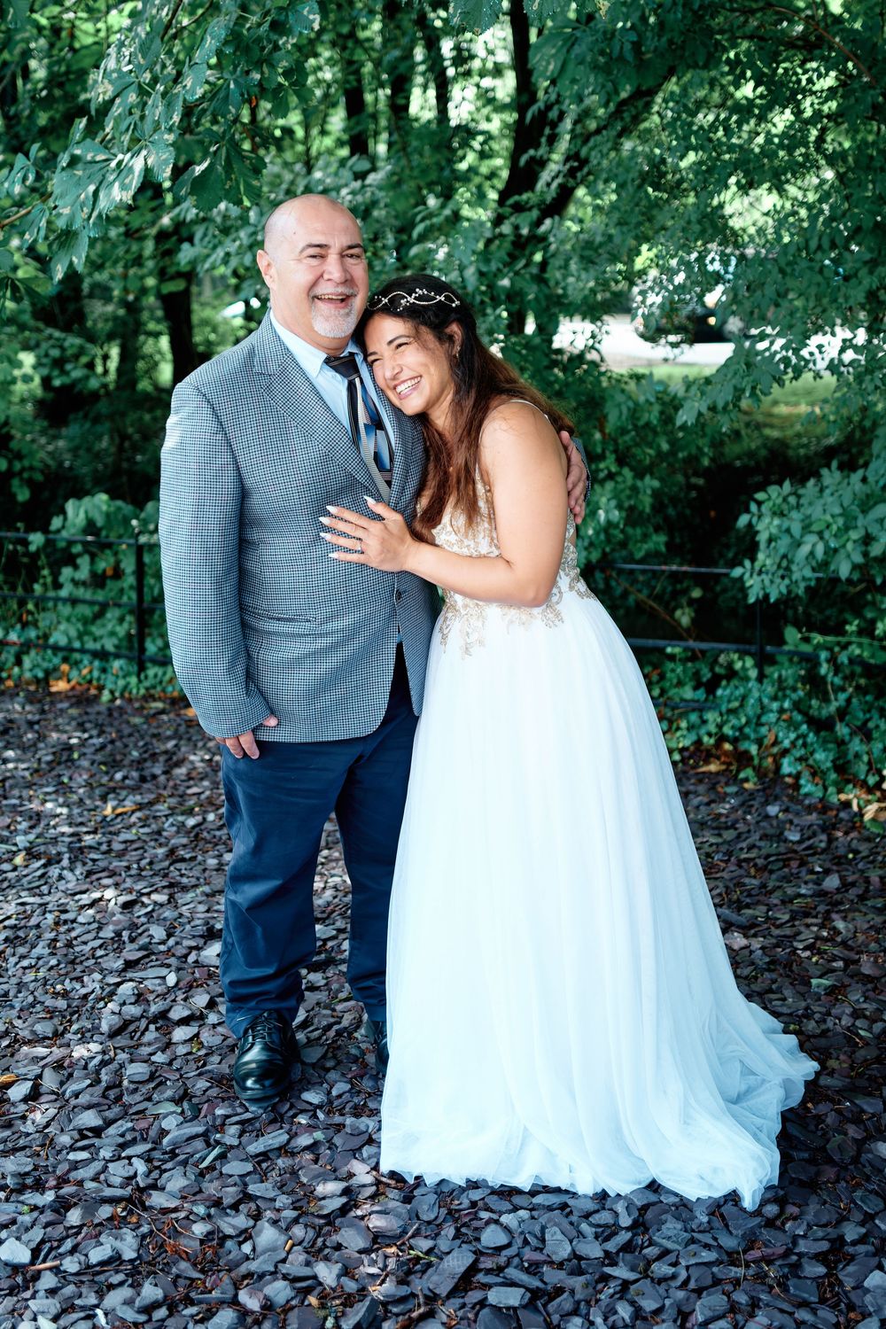 Bride in white gown and groom in gray blazer smiling together outdoors among lush green trees.