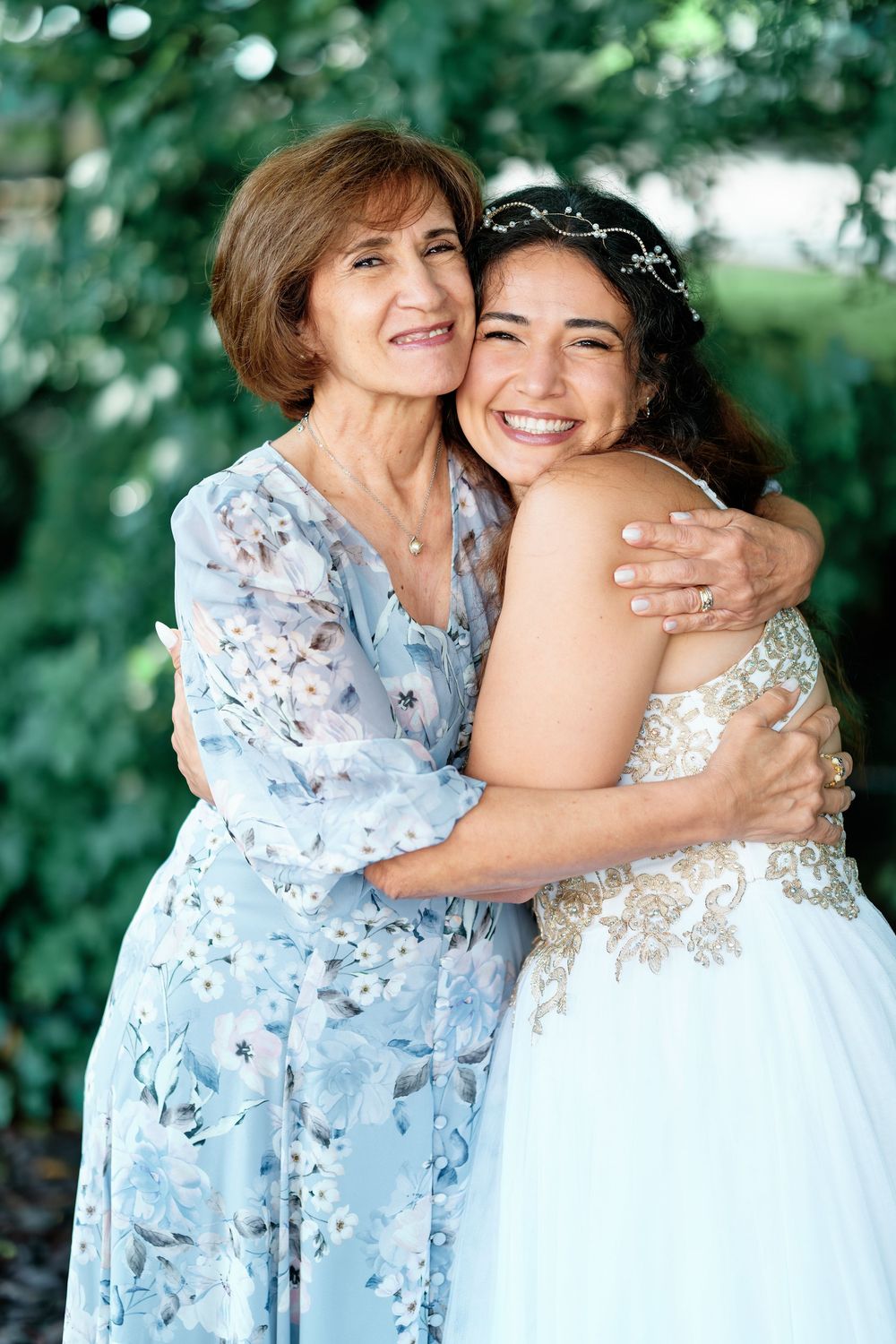 A bride in a white lace gown and floral crown hugs an older woman in a blue floral dress, smiling outdoors.