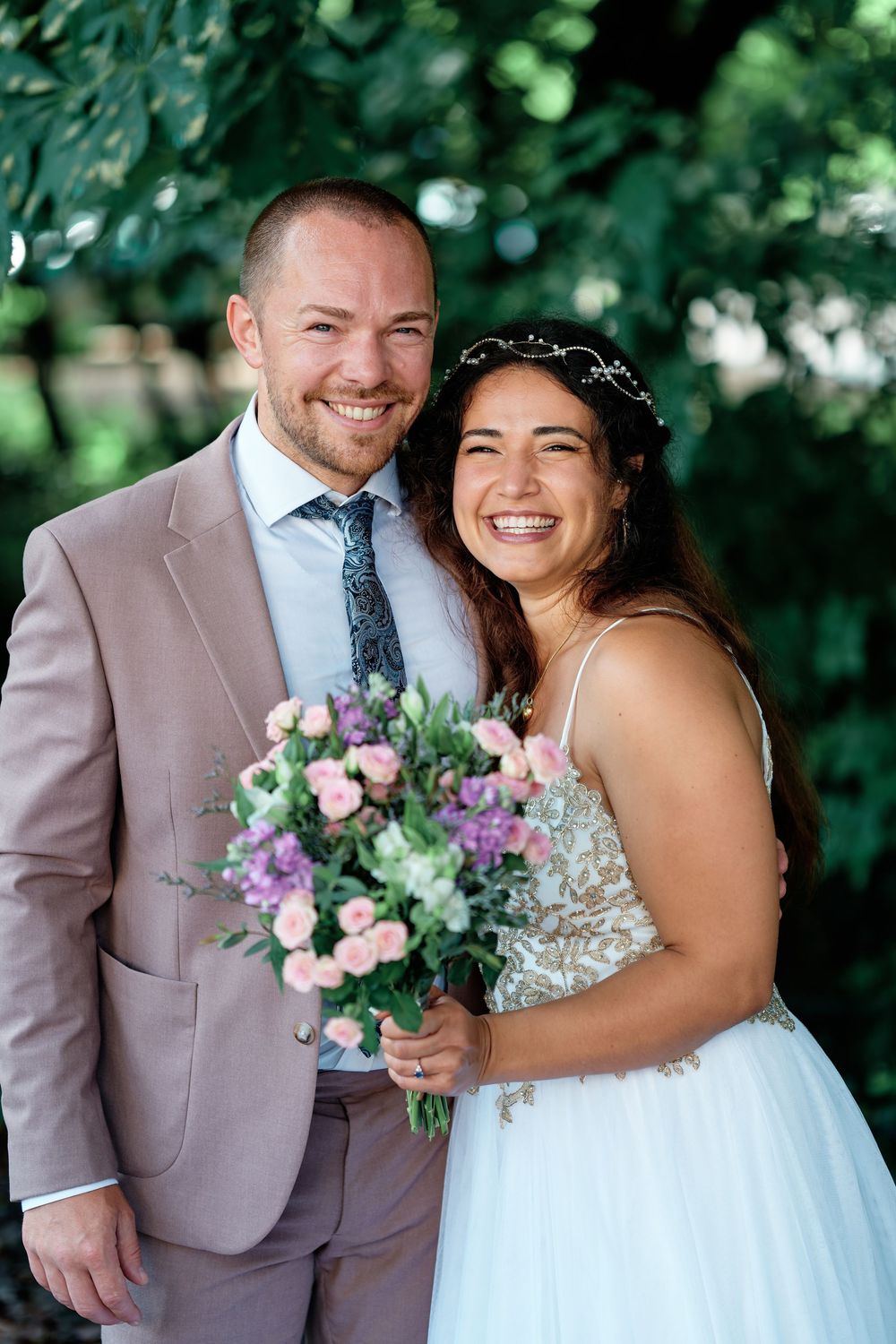 Smiling bride and groom pose outdoors, bride holding pink and purple bouquet, wearing white gown and floral crown.
