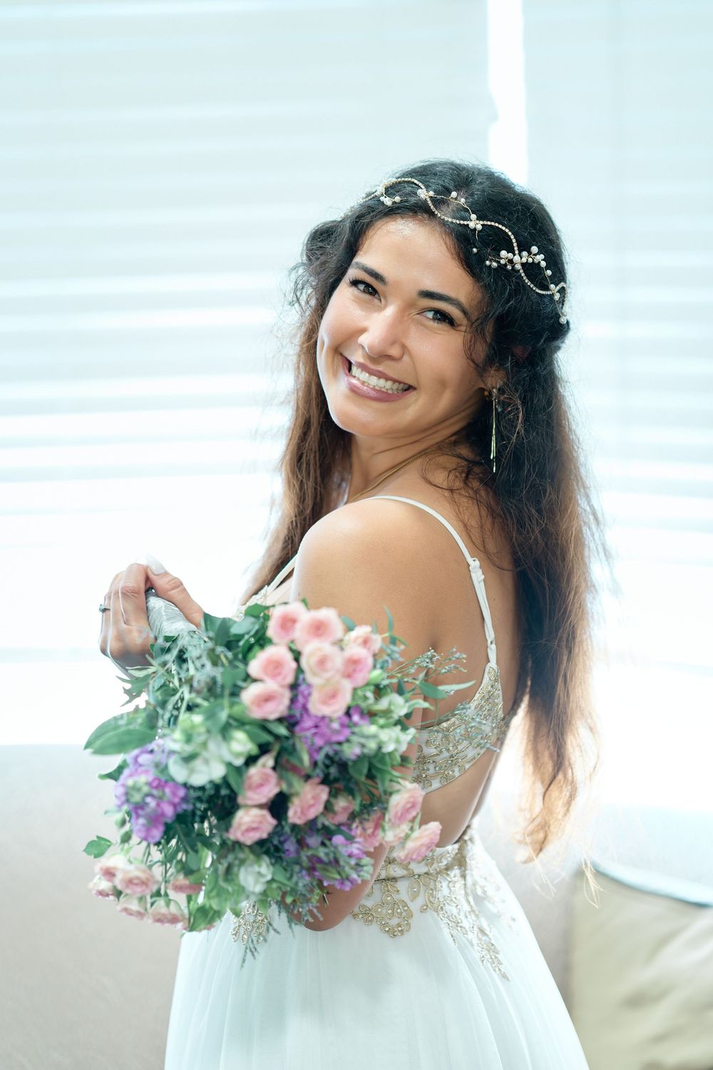 Smiling bride in white dress holding pink and purple flower bouquet with floral hair accessory.