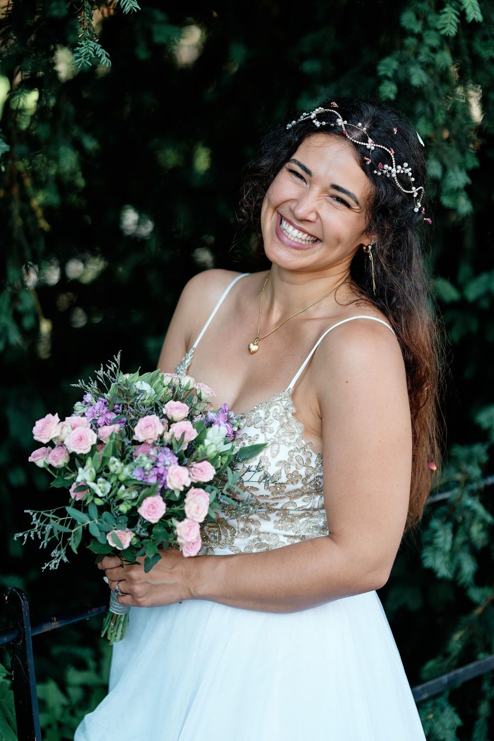 Smiling bride in white dress holds pink and purple bouquet, wearing floral crown, standing before lush green foliage.