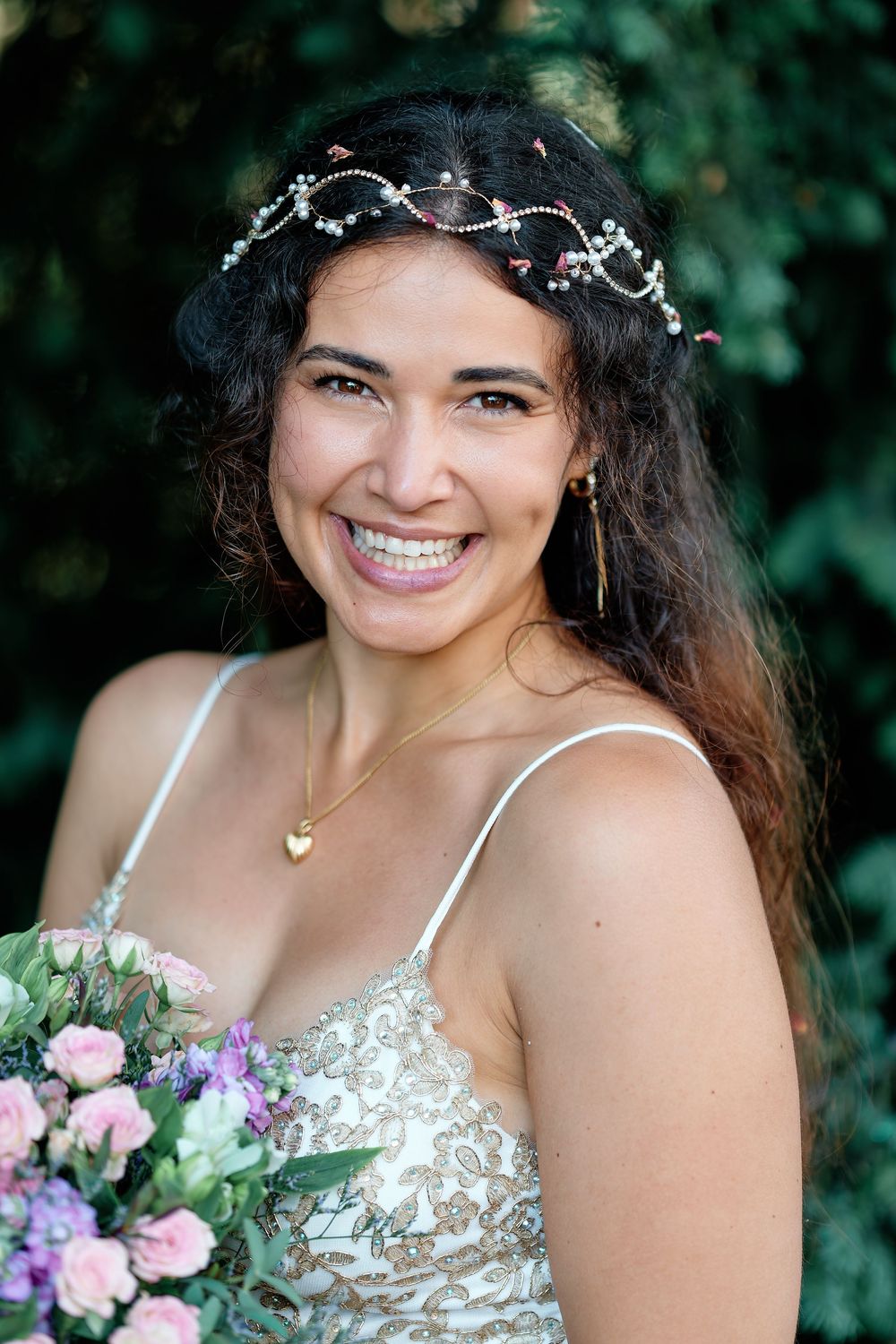 Smiling bride wearing a floral crown and lace dress holds a pink bouquet outdoors.