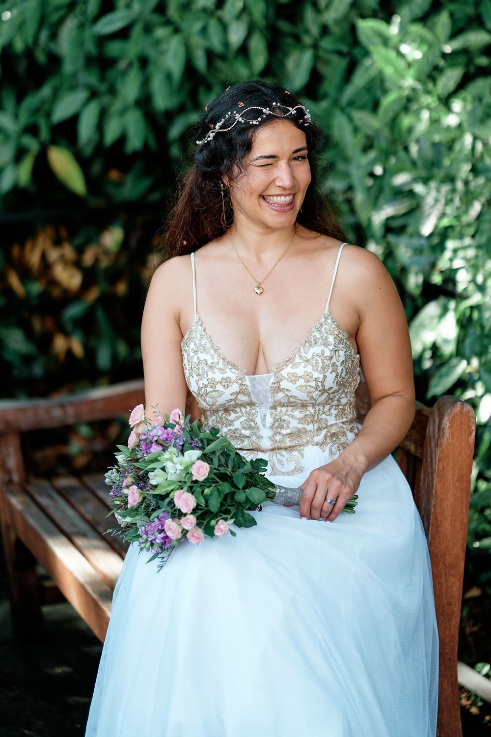 Smiling bride in gold embroidered gown holding pink bouquet, seated on wooden bench with lush green foliage backdrop.