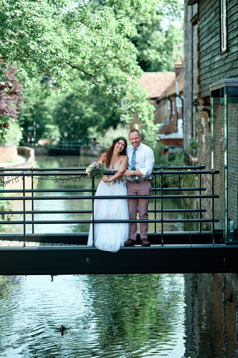 Bride and groom embracing on a bridge over a canal surrounded by lush green trees on their wedding day.