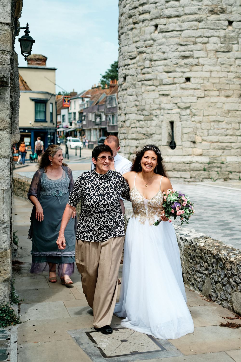 Bride in white gown holding bouquet walks with guests near historic stone tower on cobblestone street.