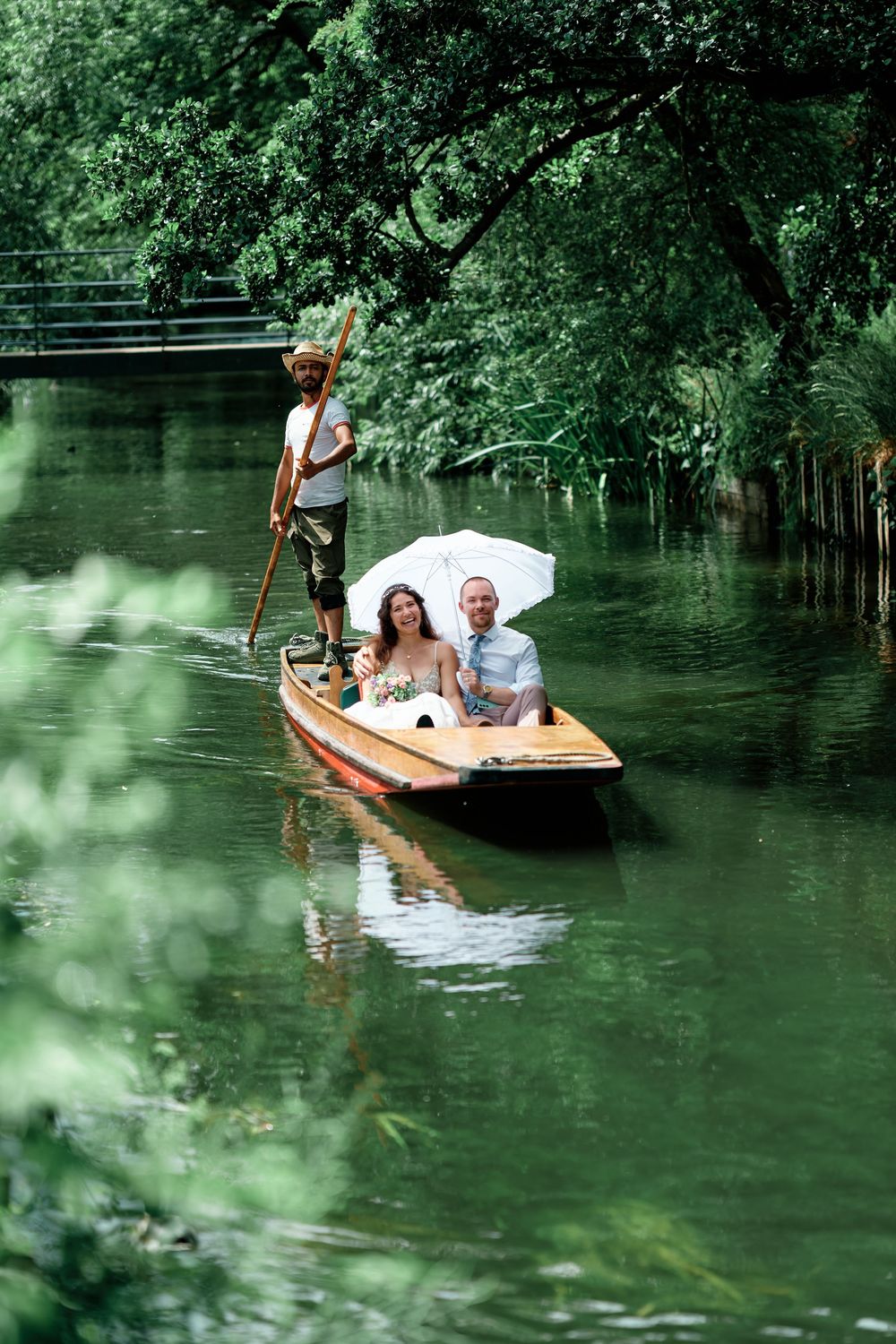 Couple in wedding attire riding a wooden punt boat on a serene green river, guided by a gondolier with a pole.