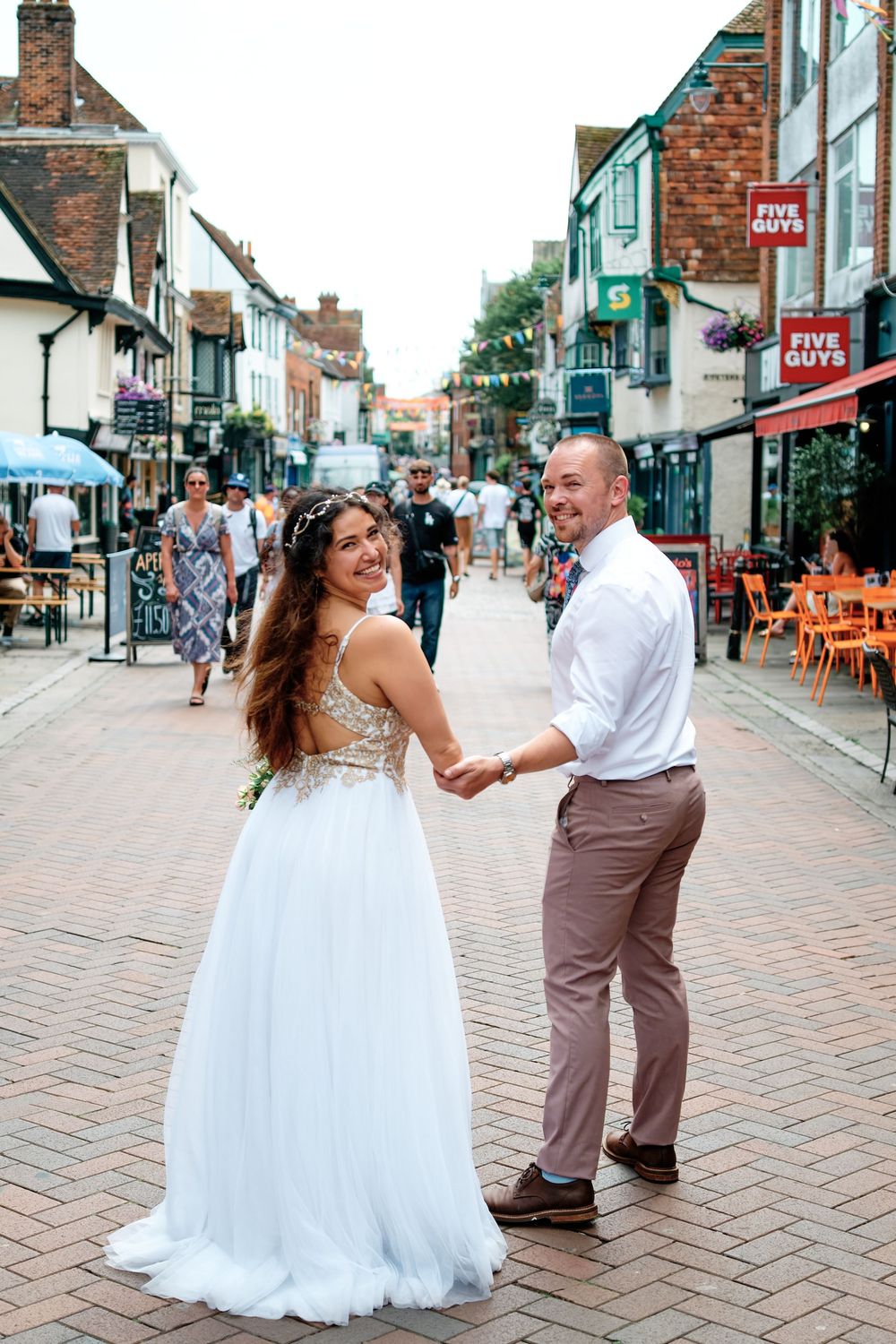 Bride and groom holding hands on a cobblestone street, smiling back at camera, with Five Guys restaurant in background.