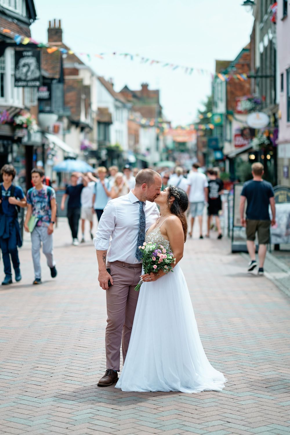 Bride and groom kissing on a busy cobblestone street, bride holding a floral bouquet in a white wedding dress.