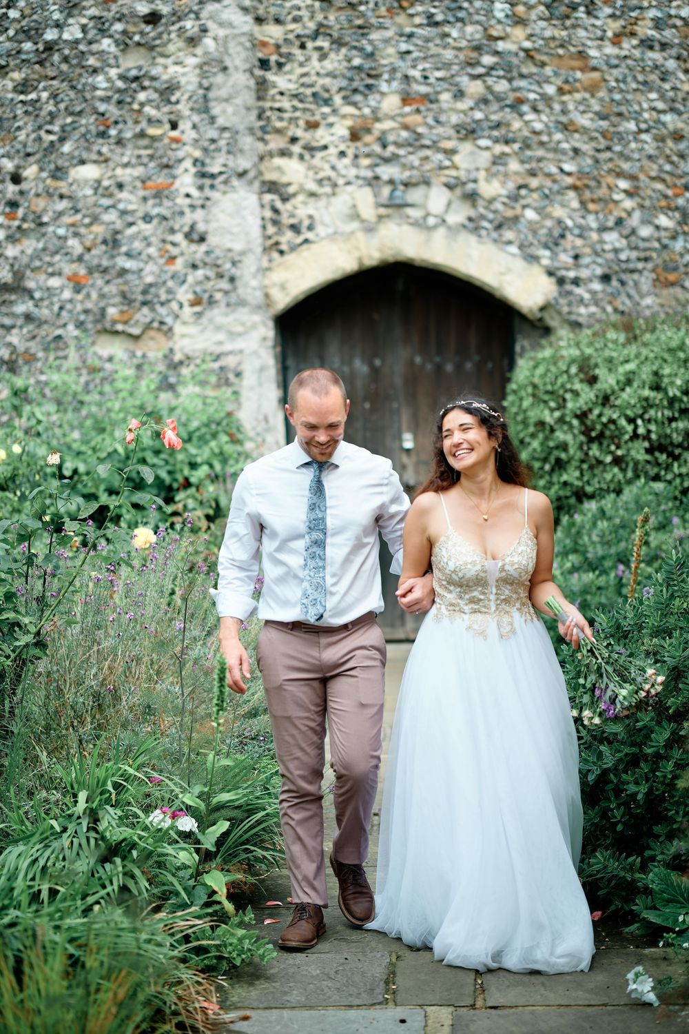 A smiling bride in a floral lace gown walks with her groom through a lush garden beside a stone wall.