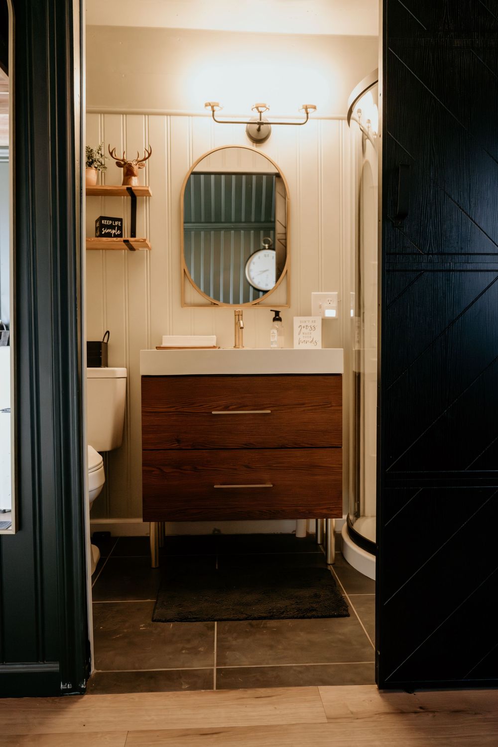 Modern bathroom with wood vanity, oval mirror, gold fixtures, beadboard walls, and dark barn door entrance.