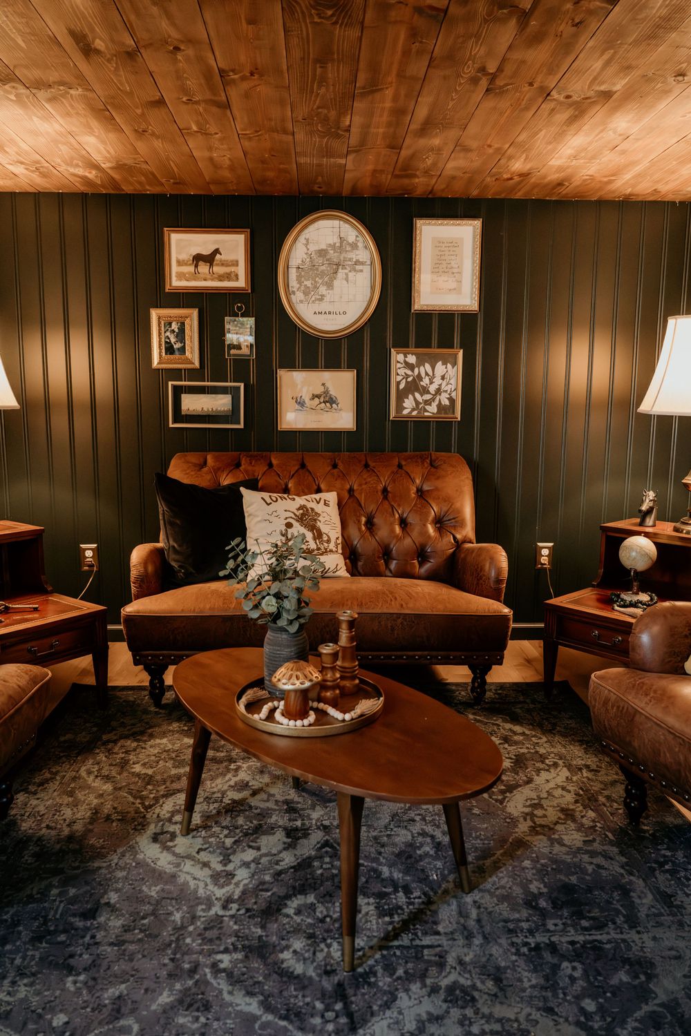 Cozy vintage living room with tufted brown leather sofa, wood ceiling, dark green walls, gallery art, and oval coffee table.