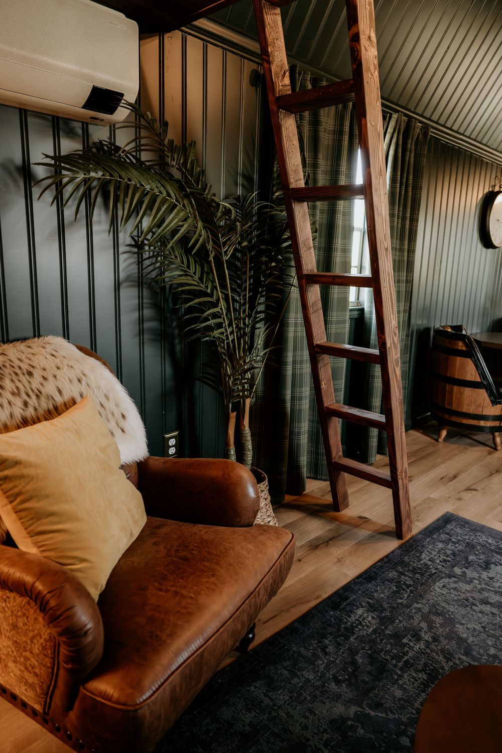 Rustic cabin interior with brown leather sofa, wooden ladder, palm plant, and dark green paneled walls on hardwood floors.