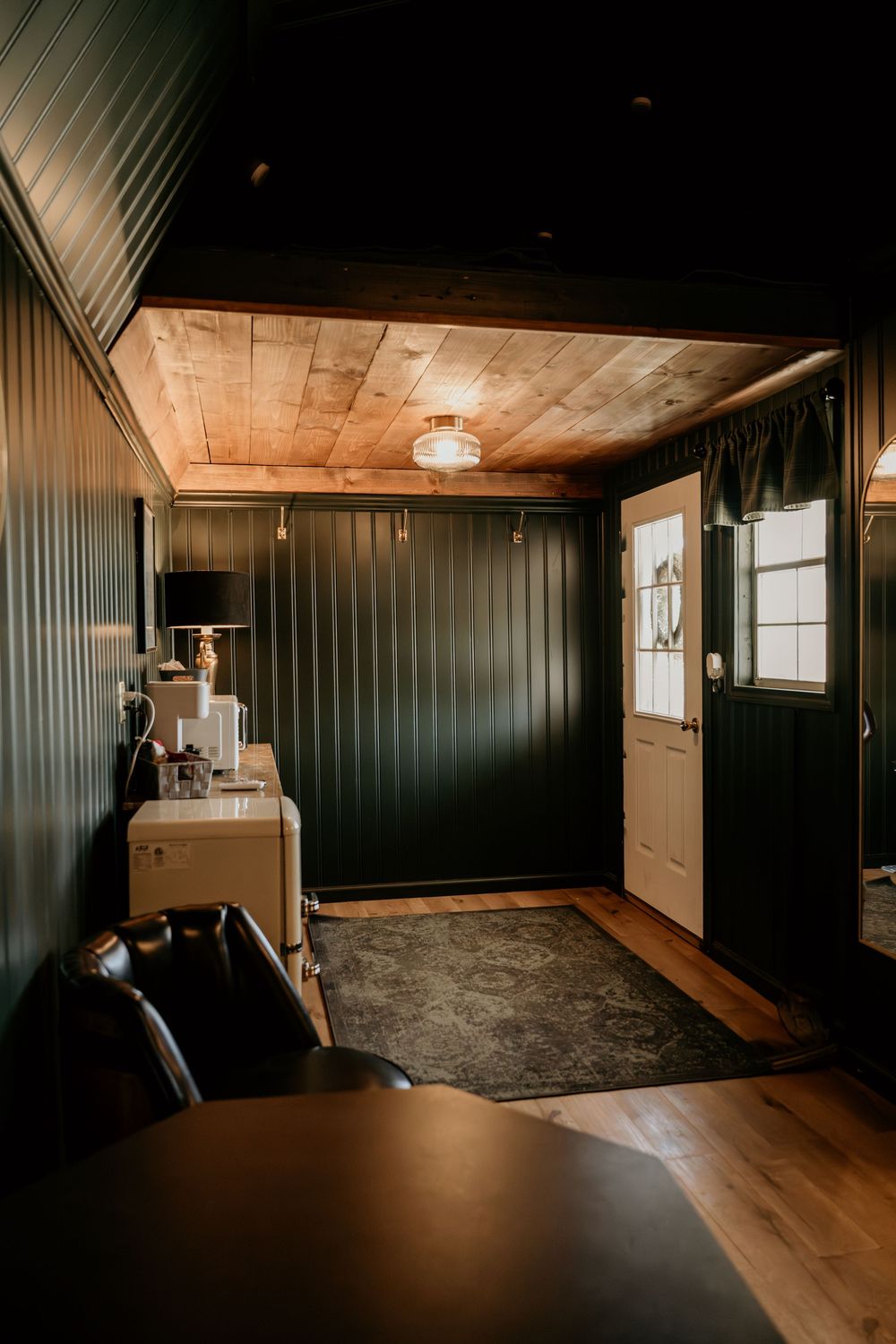 Cozy cabin interior with dark green shiplap walls, wood ceiling, hardwood floors, and a white door with natural light.