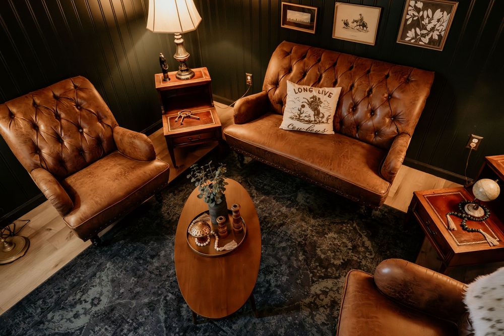 Vintage brown tufted leather loveseat and armchair with oval coffee table on blue rug in dark elegant living room.
