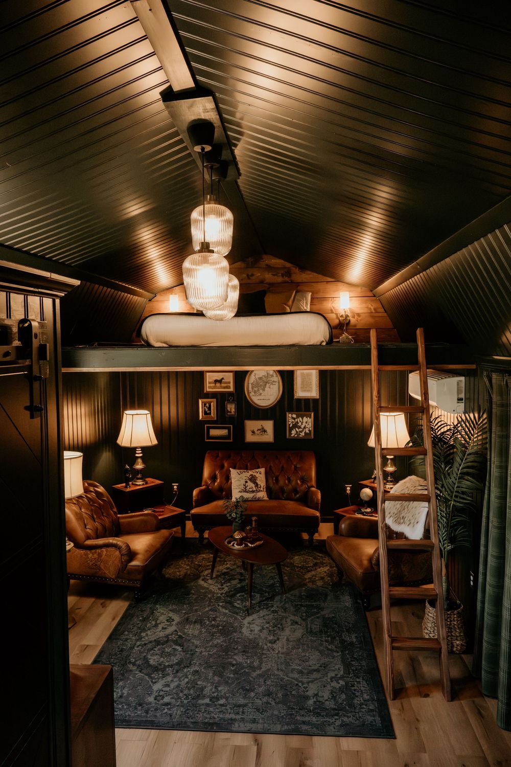 Cozy cabin interior with leather chairs, loft bed, warm pendant lights, dark wood paneling, and a vintage rug.