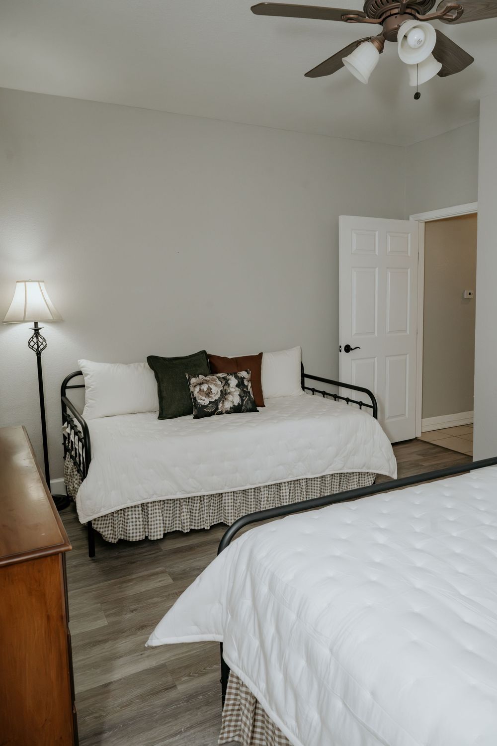 Cozy bedroom with black iron daybed, white bedding, decorative pillows, hardwood floors, ceiling fan, and warm lamp lighting.