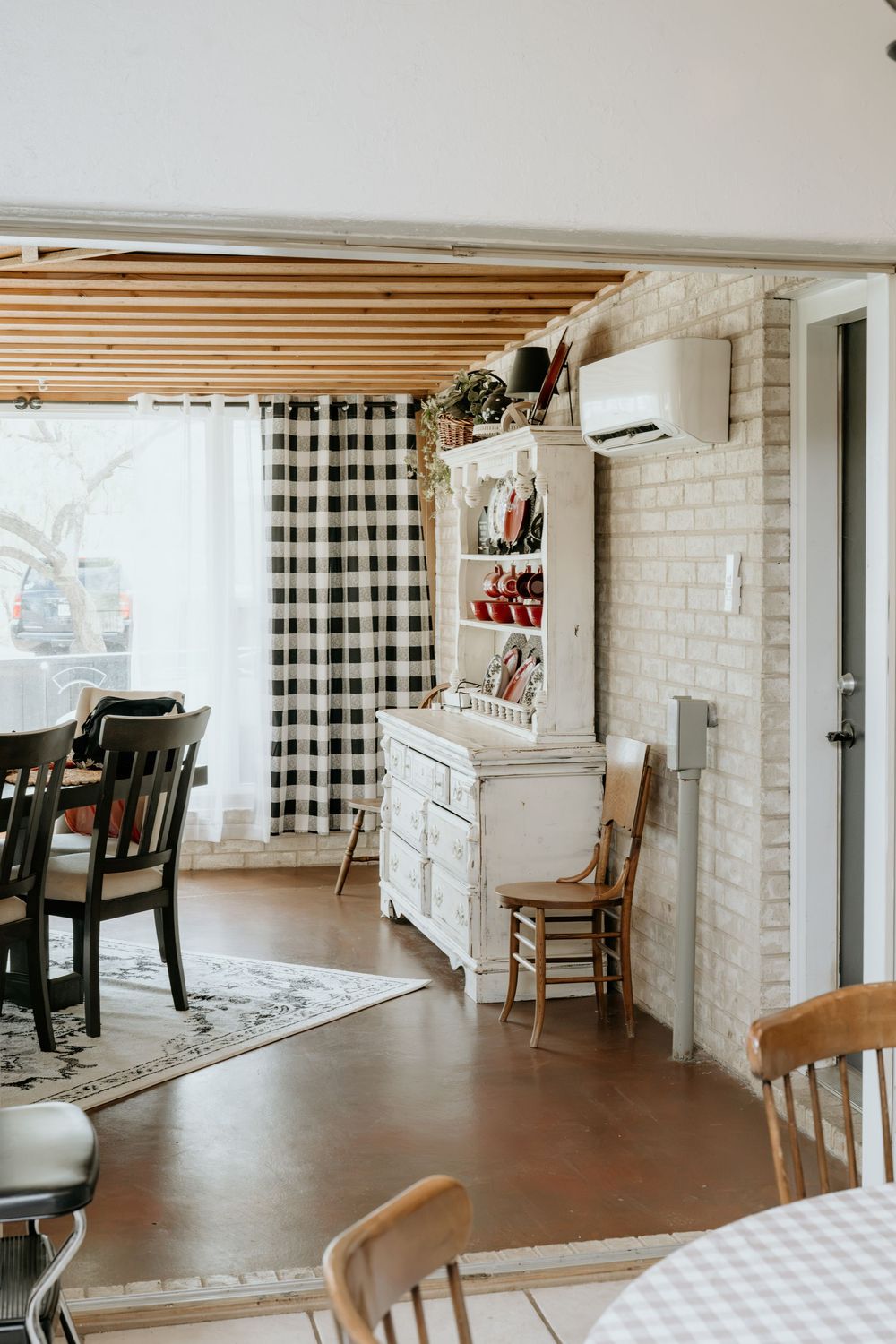 Rustic farmhouse dining area with buffalo check curtains, whitewashed brick wall, distressed cabinet, and wooden ceiling beams.