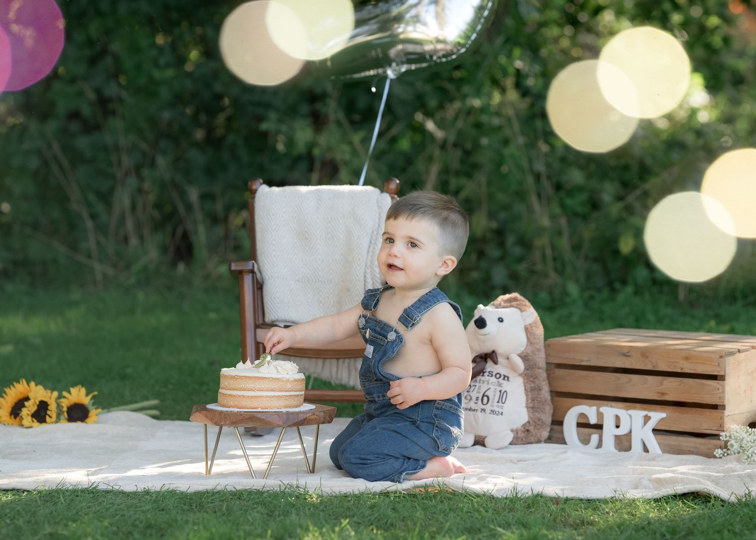 Baby boy in denim overalls sits outdoors at his first birthday cake smash with a teddy bear and balloon.
