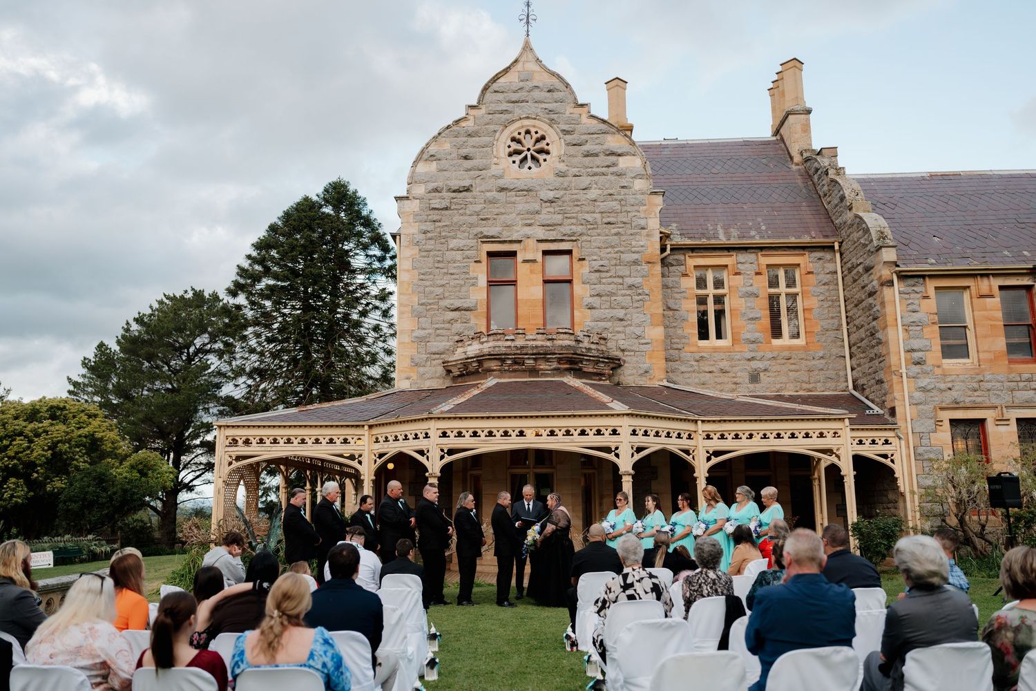 Outdoor wedding ceremony on lush lawn before historic stone manor with clock tower and covered veranda.