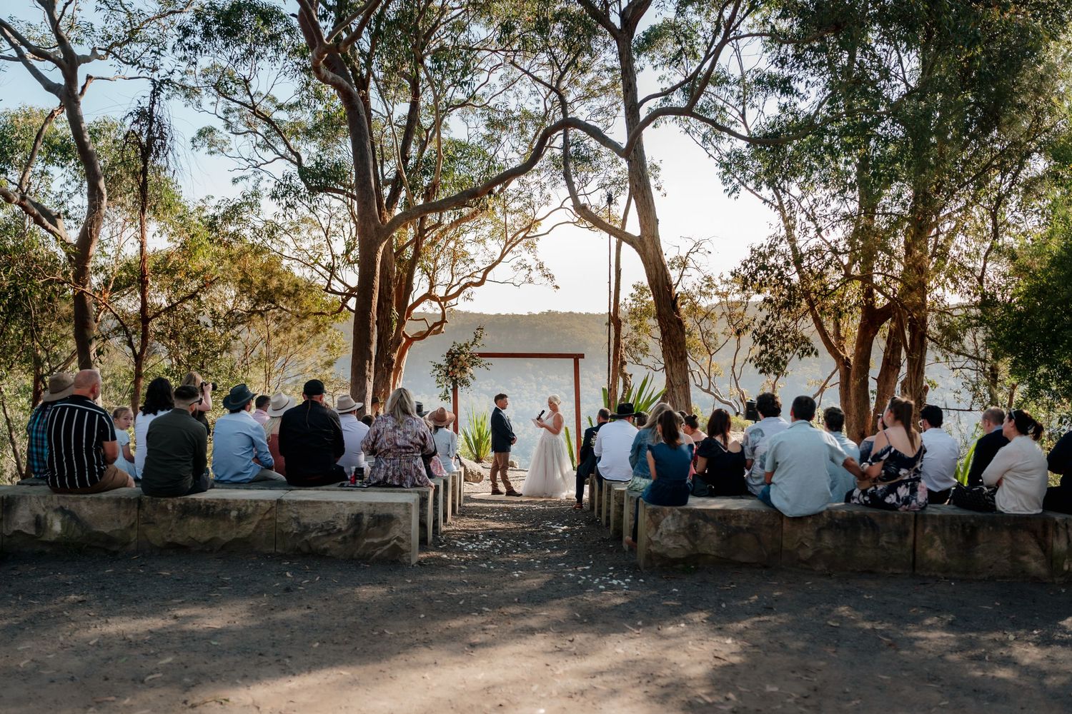 Outdoor wedding ceremony under eucalyptus trees with guests seated on stone benches facing couple at wooden arch altar.