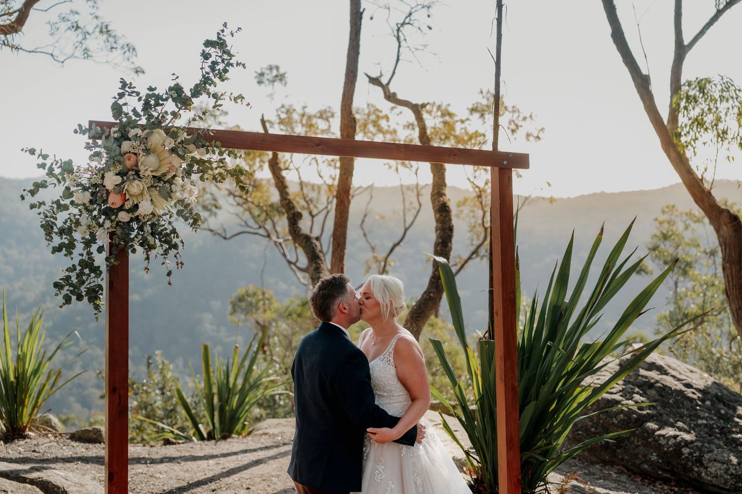 Couple shares first kiss under rustic wooden arch with floral decor, overlooking scenic mountain valley at outdoor wedding.