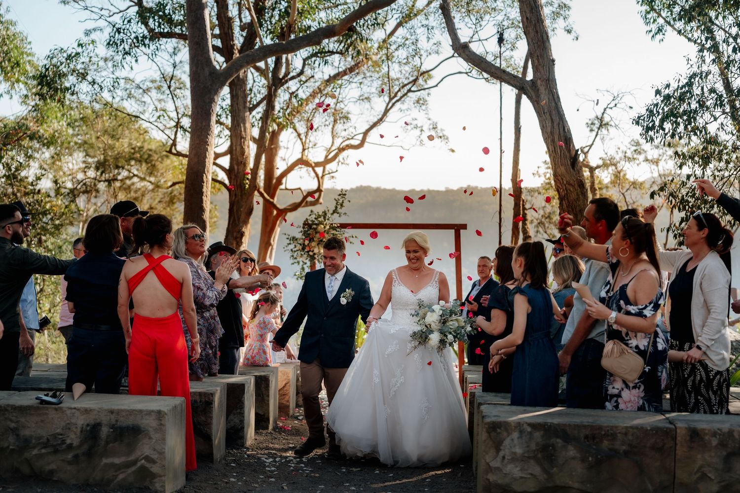 Bride and groom walk down the aisle as guests toss rose petals at an outdoor wedding ceremony.
